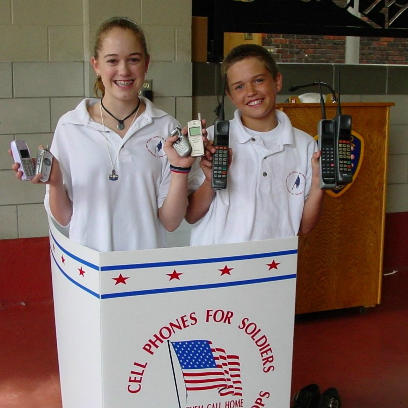 Young Brittany and Robbie Bergquist smiling behind a Cell Phones for Soldiers booth, holding various mobile phones, promoting donations to support troops.